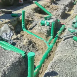 Wykop z ułożonymi zielonymi rurami kanalizacyjnymi przed zasypaniem, widoczny fragment ciężarówki z boku.