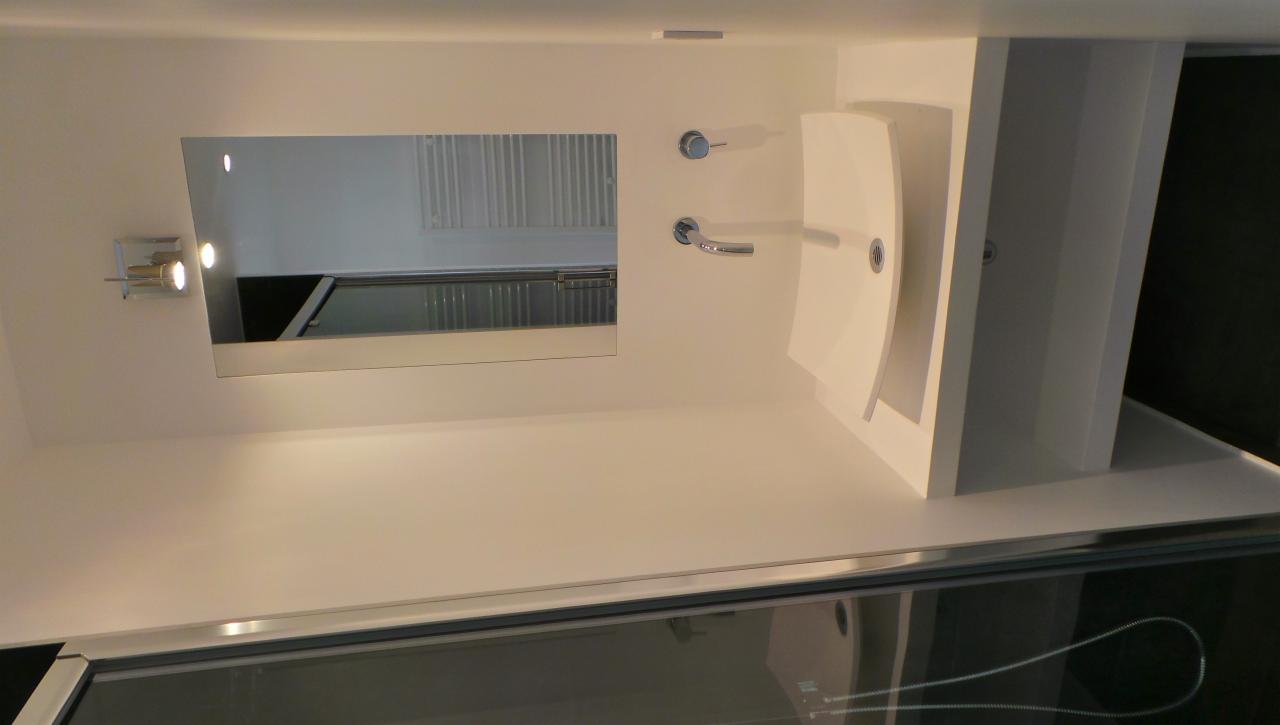 Nowoczesna, minimalistyczna łazienka z białą umywalką o zaokrąglonym kształcie, wbudowaną w blat, oraz lustrem z oświetleniem punktowym, odbijającym grzejnik drabinkowy.