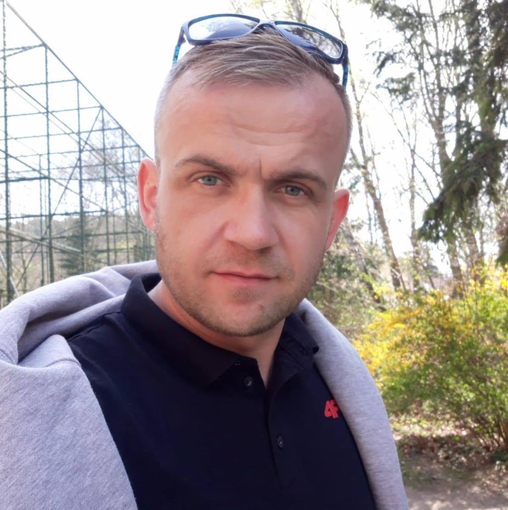 Mężczyzna z niebieskimi oczami w czarnej koszulce polo i szarej bluzie z kapturem, okulary na głowie, tło z drzewami i metalową konstrukcją.