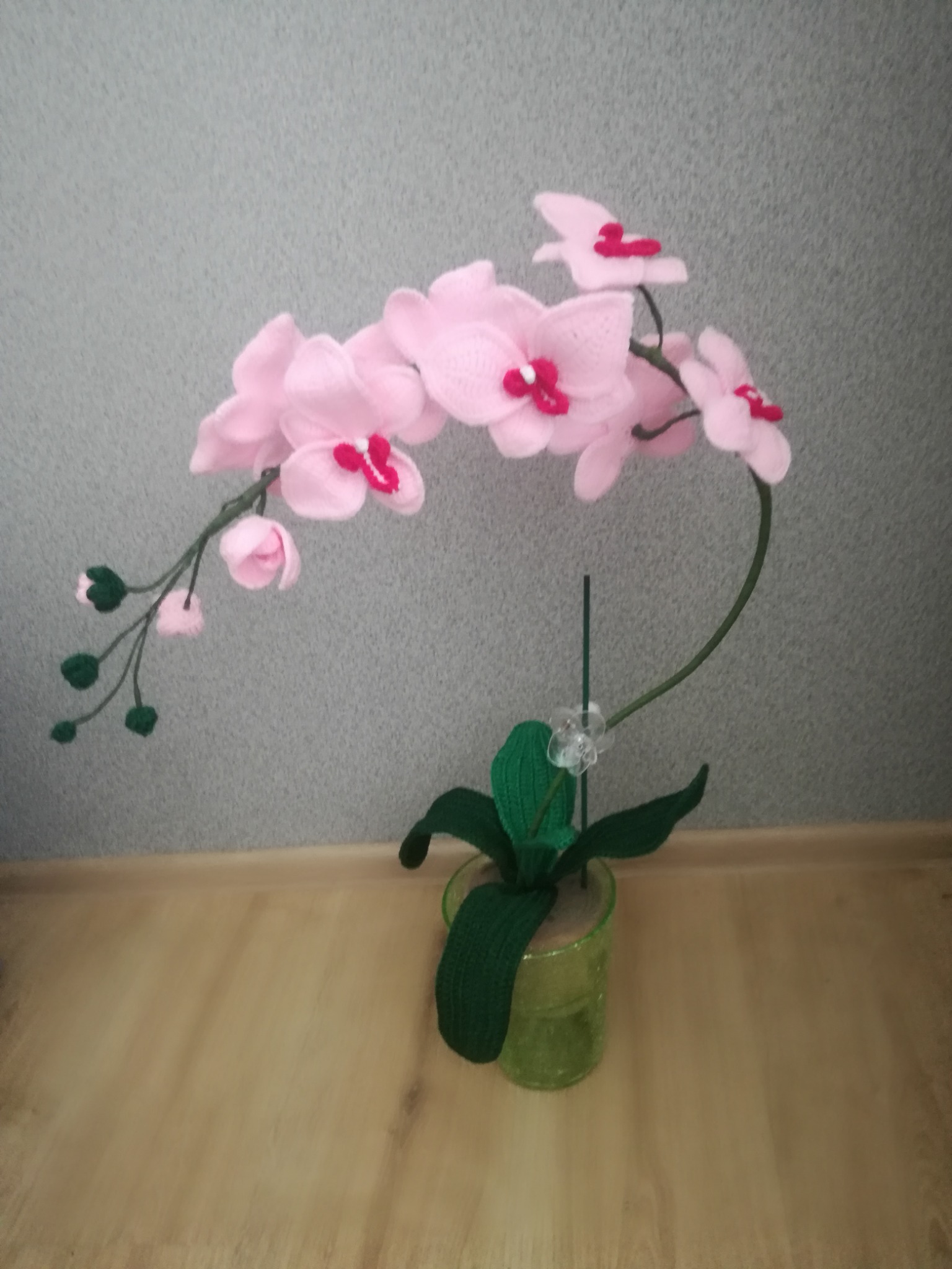 Ręcznie robiona, dziergana na drutach różowa orchidea w zielonym, szklanym wazonie, stojąca na drewnianej podłodze na tle szarej ściany.