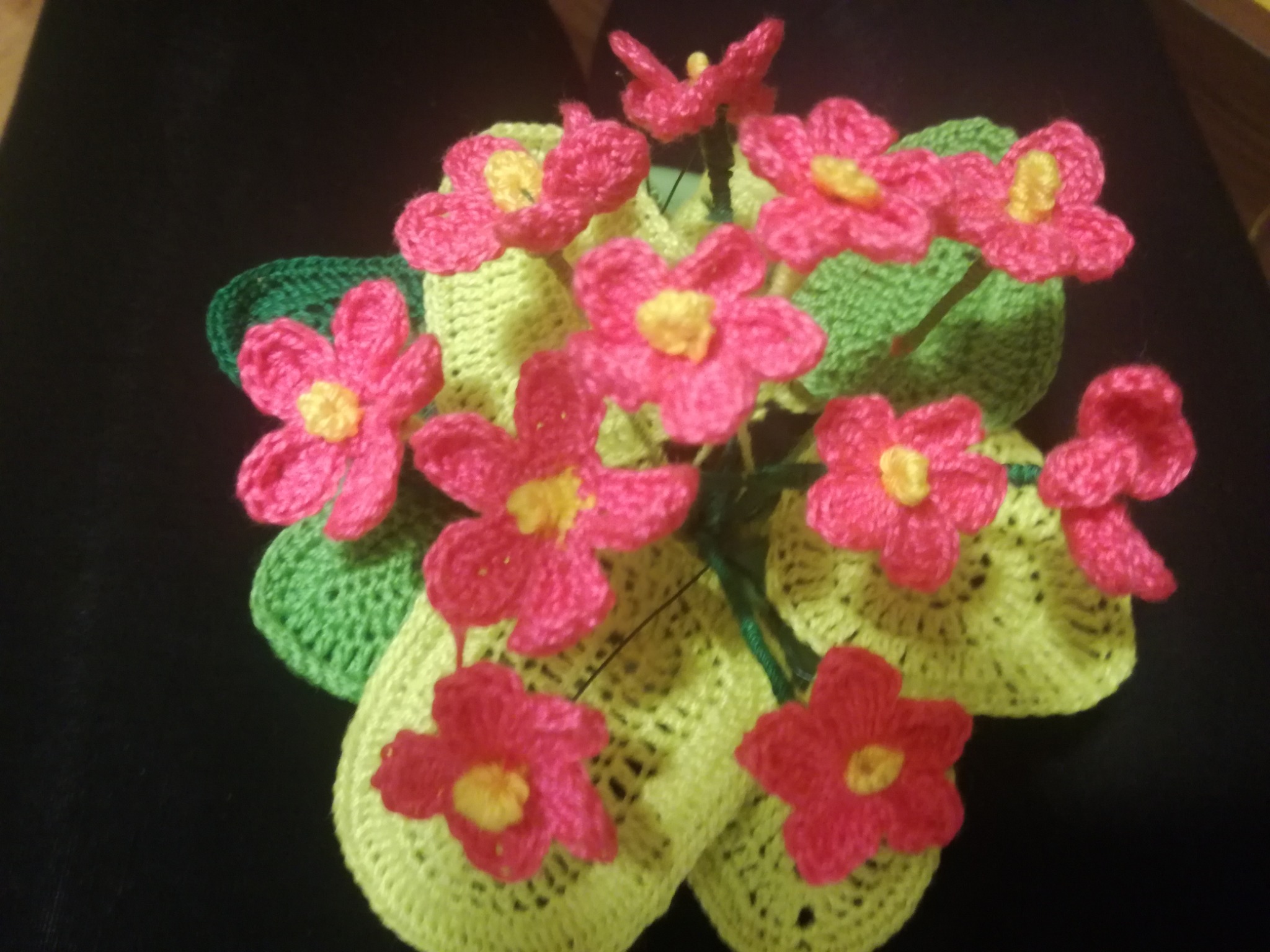 Ręcznie robiony bukiet z dzierganych kwiatów w odcieniach różu i zieleni na ciemnym tle, z widoczną fakturą włóczki.