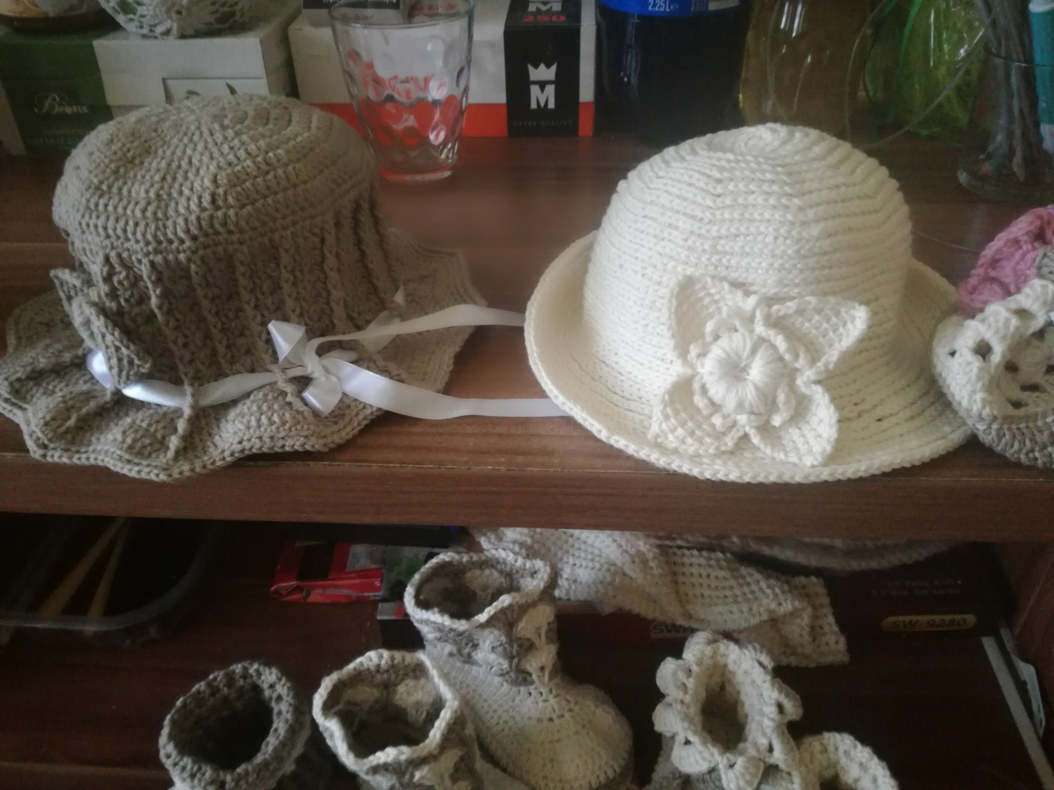 Beżowy i biały, ręcznie robiony, dziewiarski kapelusz dla dziecka oraz buciki, na drewnianej półce z różnymi przedmiotami w tle.