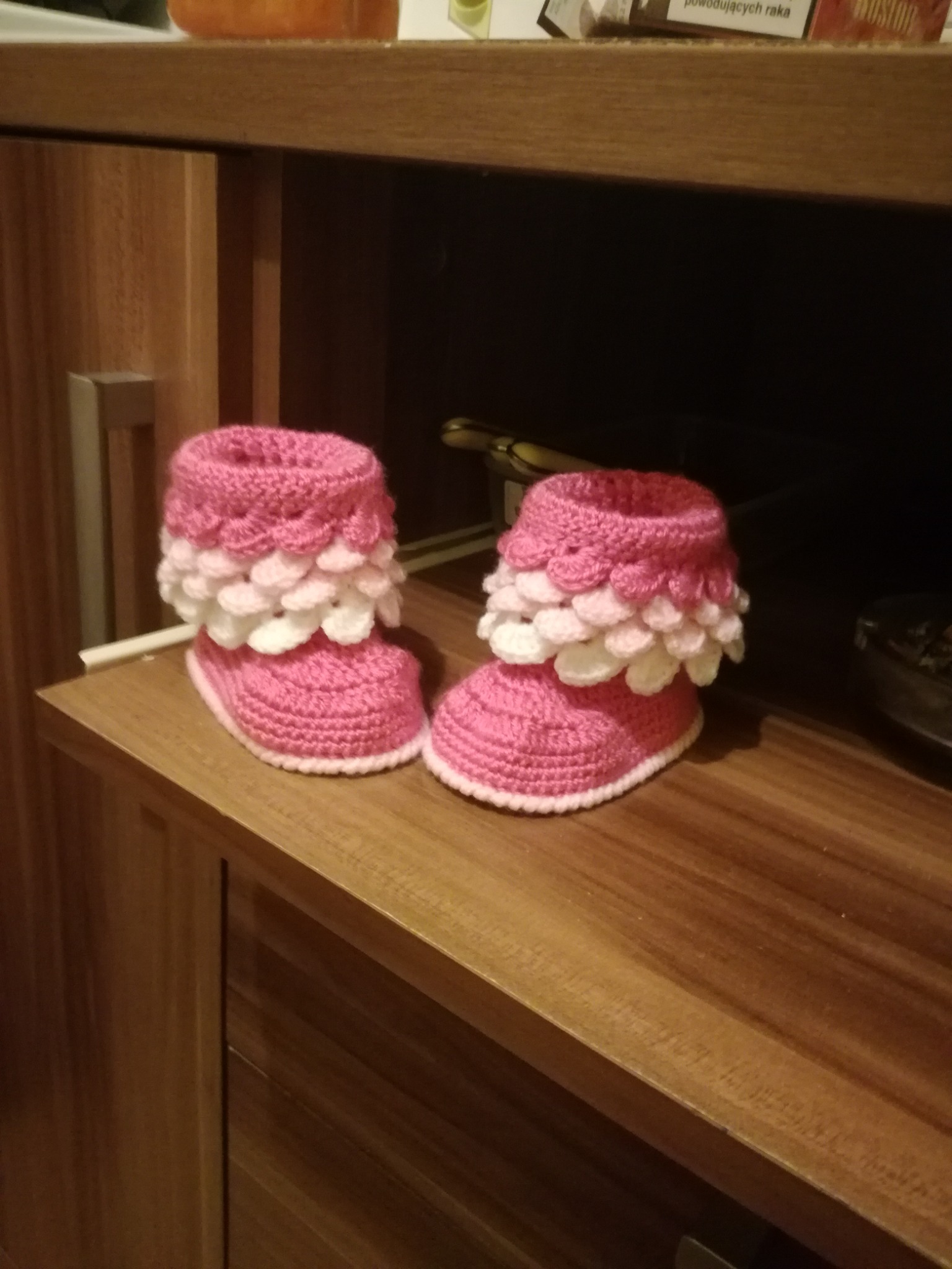 Ręcznie robione, różowe, dziergane buciki niemowlęce z białymi, falowanymi wykończeniami, ustawione na drewnianej półce.