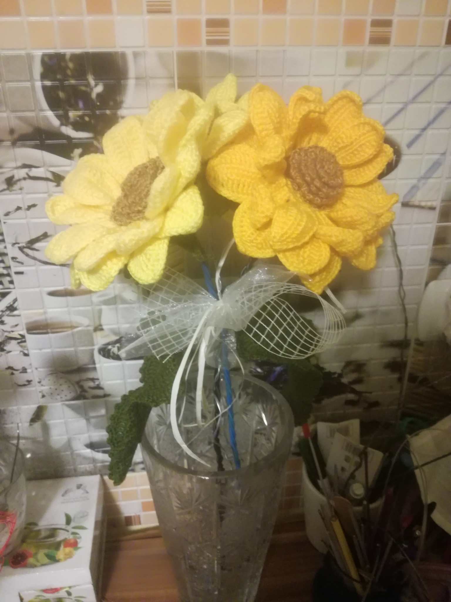 Dwa żółte, ręcznie robione, szydełkowe słoneczniki w szklanym wazonie, ozdobione białą kokardką z siatki, na tle mozaiki ceramicznej.