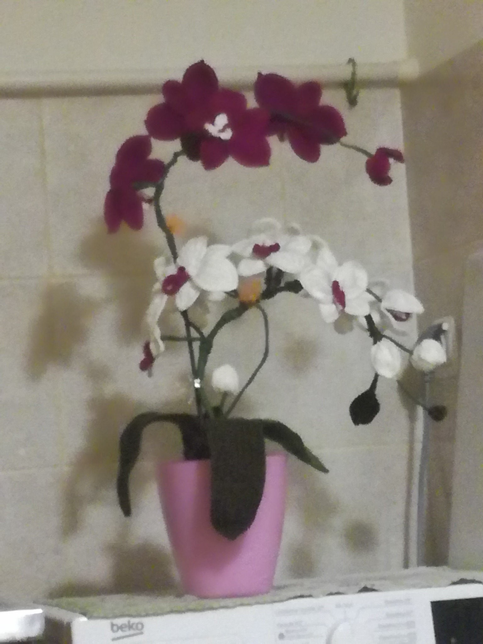 Ręcznie robiona, filcowa orchidea w różowej doniczce, stojąca na pralce Beko, z kwiatami w kolorze białym i bordowym.