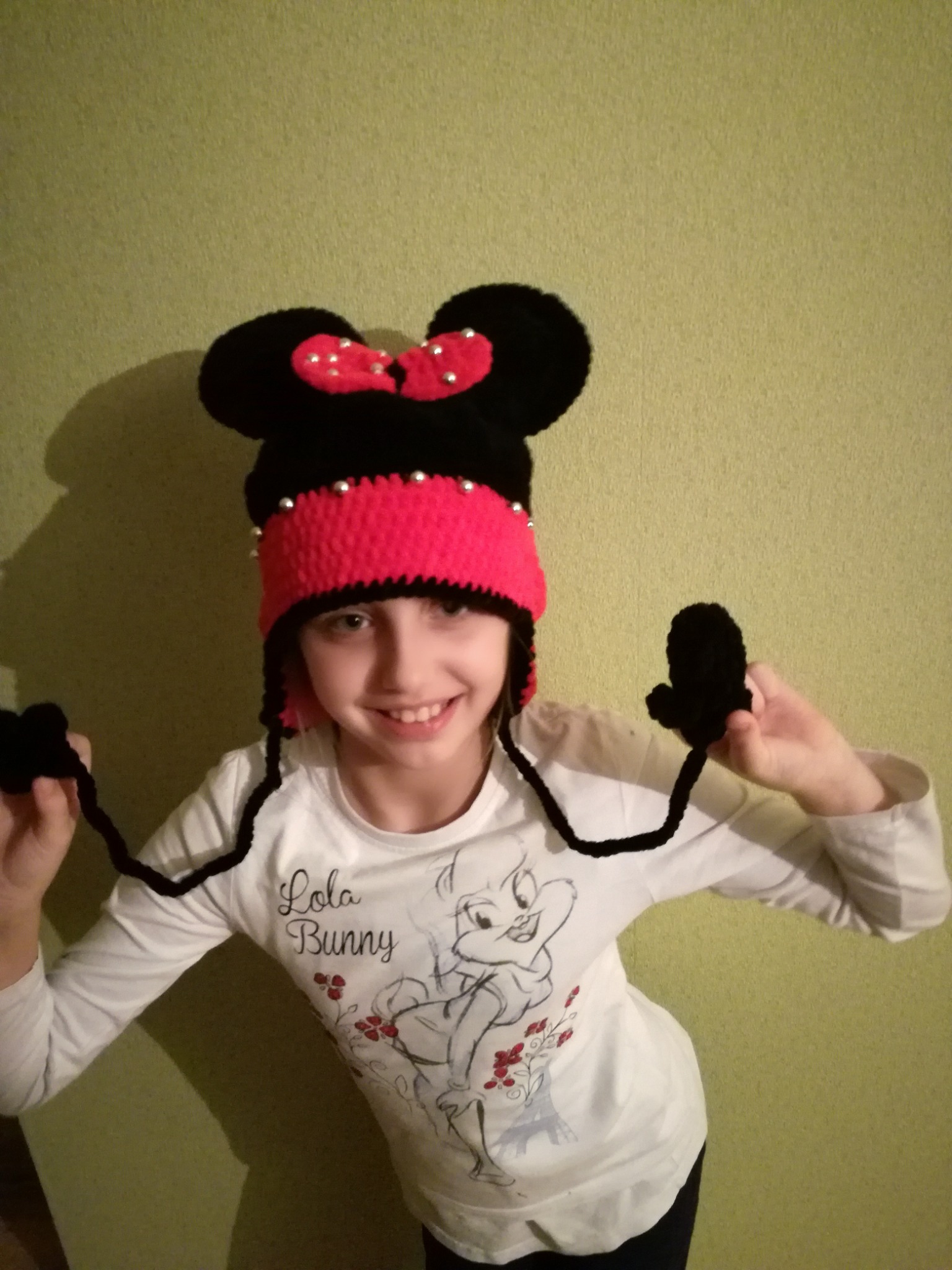 Uśmiechnięta dziewczynka w czapce z uszami Myszki Miki, ręcznie robiona z czarnej włóczki z różowym paskiem i kokardą, trzyma czarne, dziergane mitenki na sznurkach.