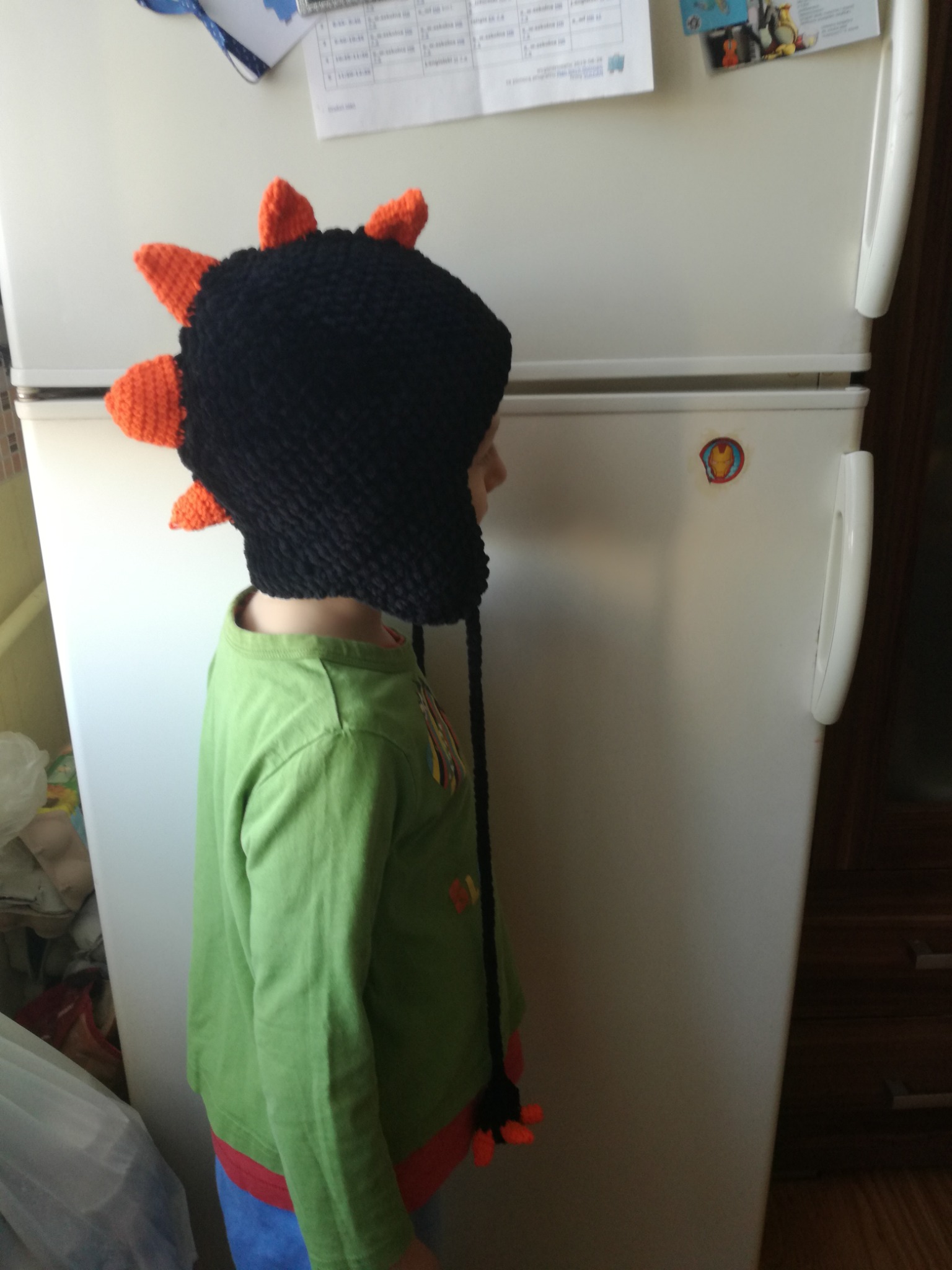 Chłopiec w czarno-pomarańczowej, dzierganej czapce w kształcie dinozaura, stojący bokiem na tle białej lodówki z naklejką Iron Mana.