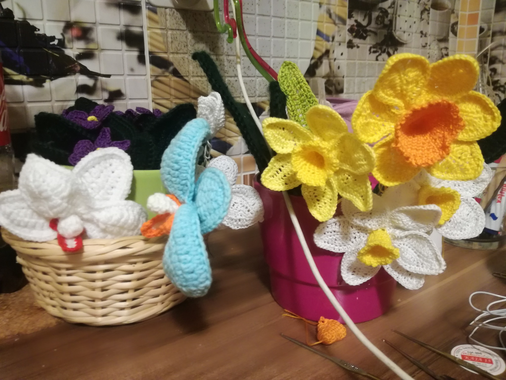 Ręcznie robione, szydełkowe kwiaty: żółte narcyzy z pomarańczowymi środkami, białe orchidee w wiklinowym koszyku z czerwonym akcentem, niebieski ptaszek, fioletowe fiołki, wszystko wykonane...