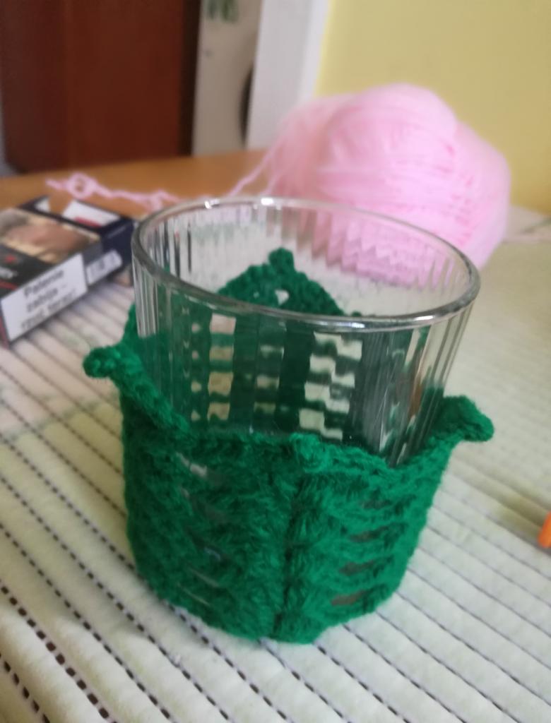Szklanka w zielonym, ażurowym, ręcznie robionym pokrowcu dziewiarskim, obok różowy kłębek włóczki i paczka papierosów na stole.