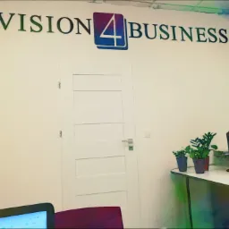 Biuro z logo 'Vision 4 Business' na ścianie, białe drzwi, rośliny doniczkowe i monitor na biurku.