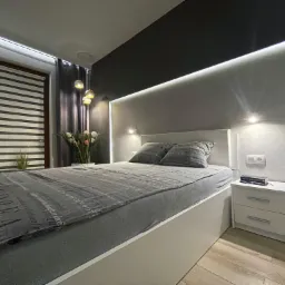 Nowoczesna sypialnia w odcieniach szarości i bieli z podświetleniem LED, drewnianą podłogą i roletami w oknie, na szafce nocnej leży książka.