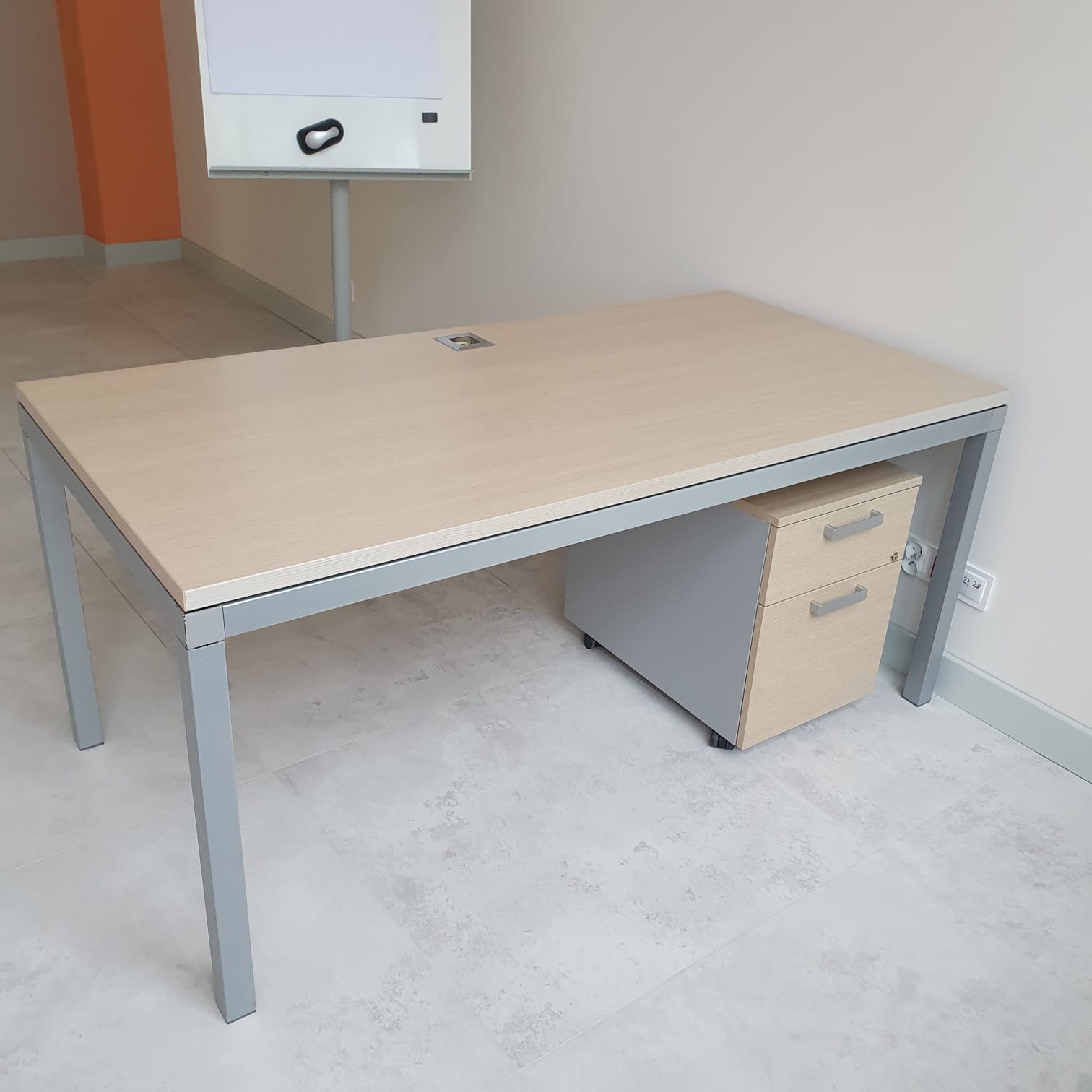 Biurko z szarym stelażem i jasnym blatem, z szafką na kółkach z dwoma szufladami, na tle białej ściany i tablicy suchościeralnej.