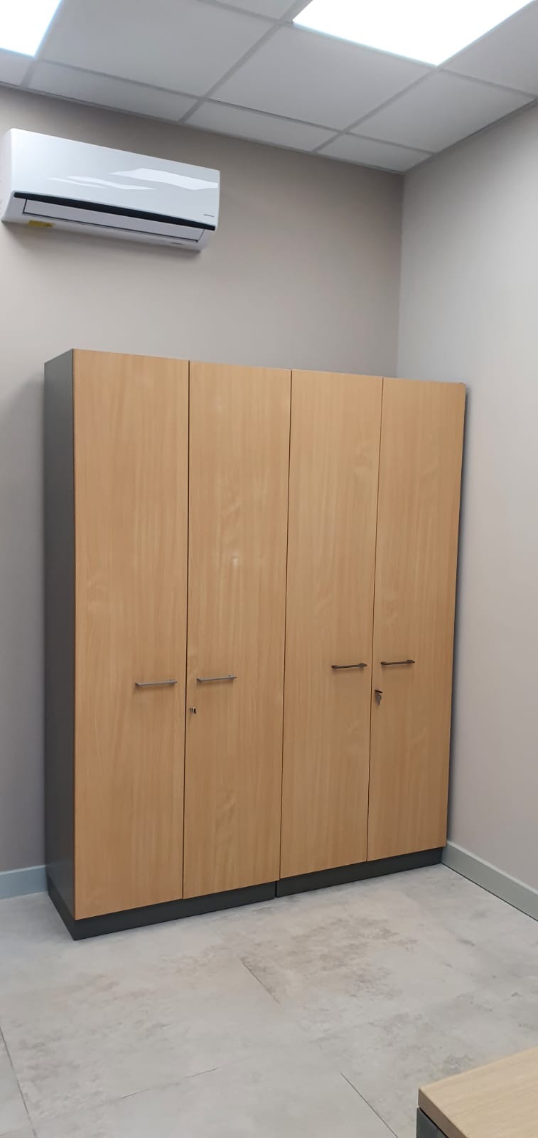 Czterodrzwiowa szafa biurowa z jasnego drewna z szarymi bokami i podstawą, z zamkiem w jednych drzwiach, na tle biurowego wnętrza z klimatyzacją na ścianie.