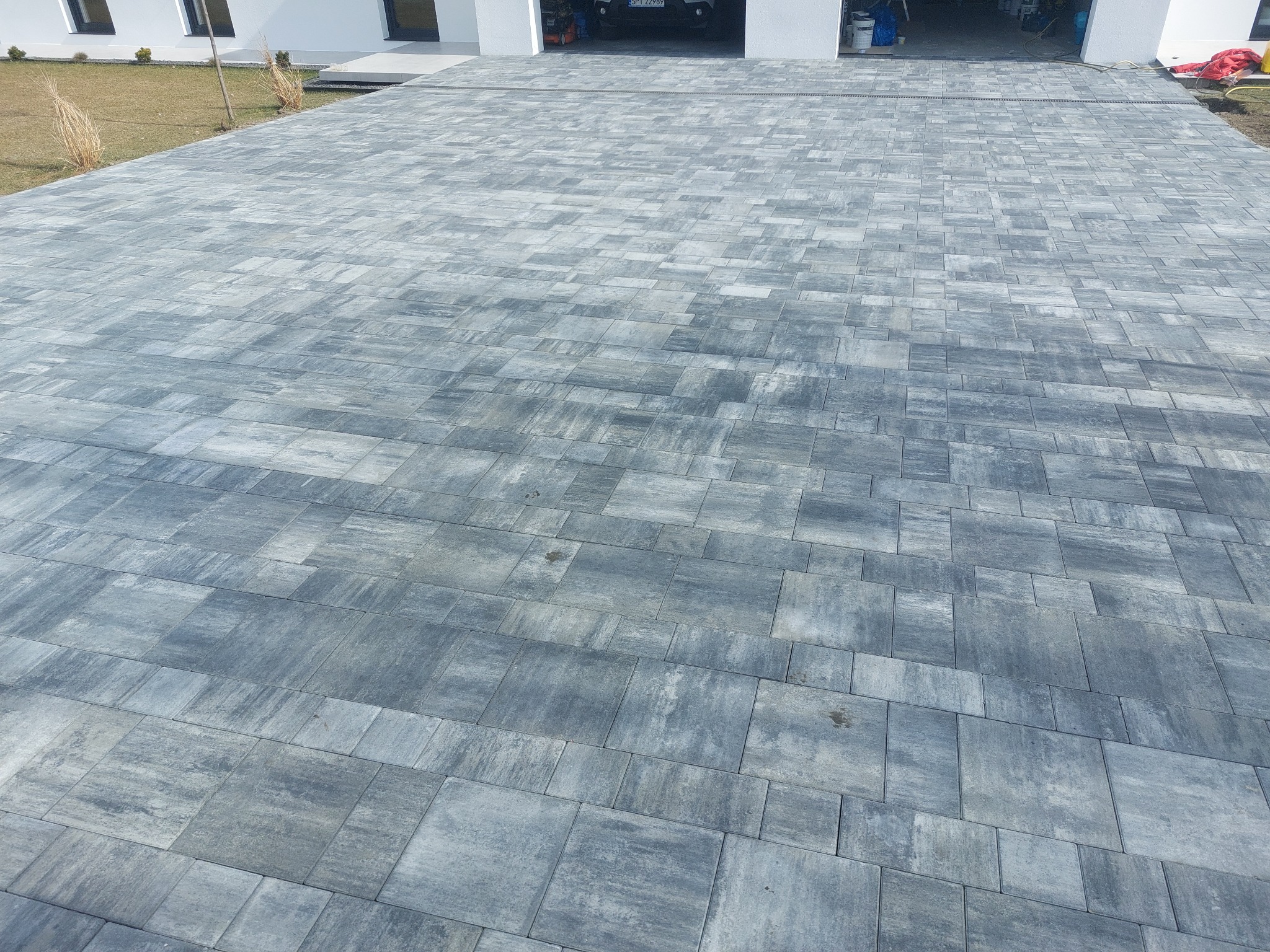 Szara kostka brukowa ułożona na podjeździe przed białym domem z garażem. Widoczna faktura kostki i wzór ułożenia. Minimalistyczny styl.