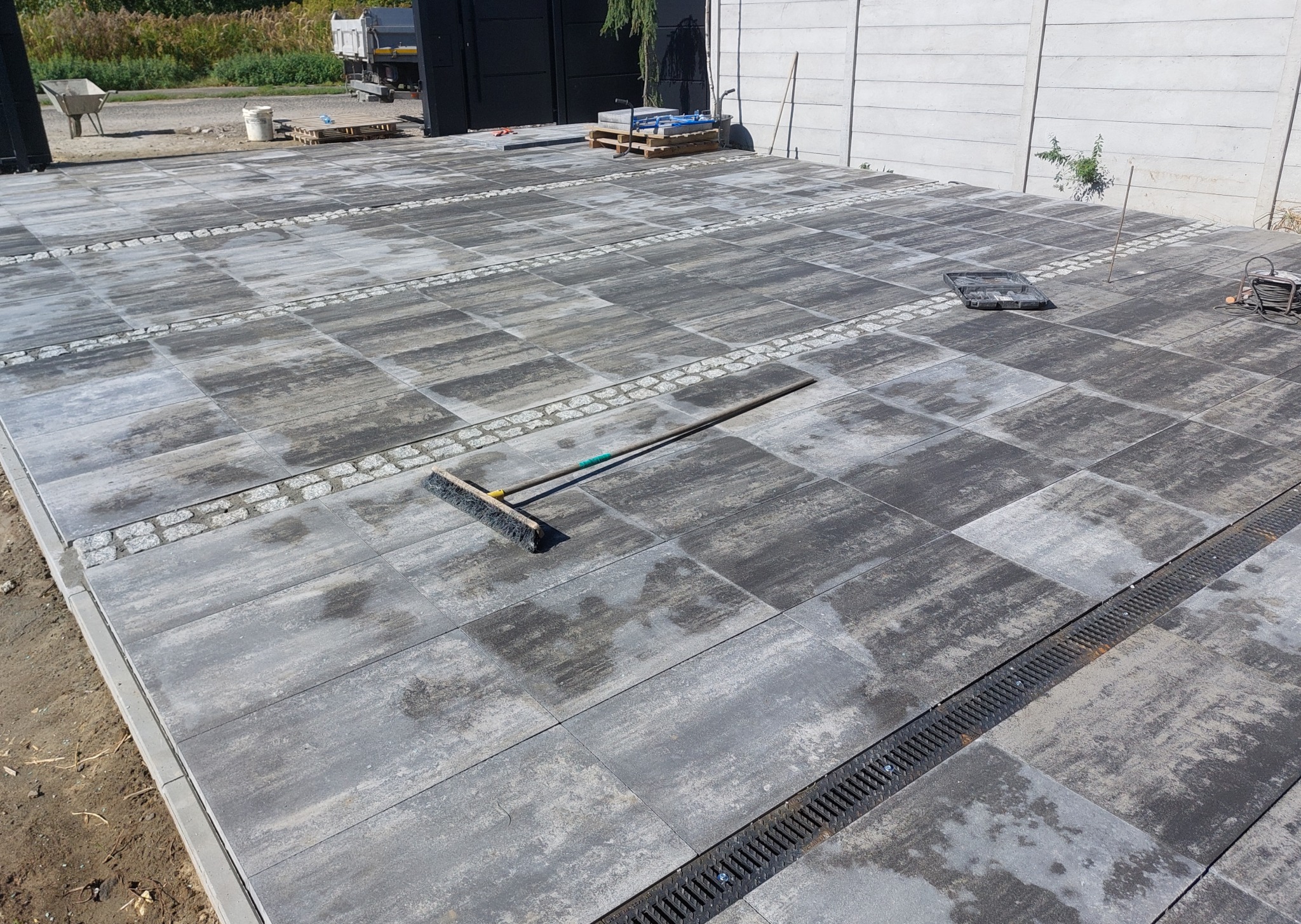 Ułożona szara kostka brukowa z dekoracyjnymi pasami z drobnych kamieni i odpływem liniowym. Widoczna szczotka brukarska i fragment ogrodzenia.