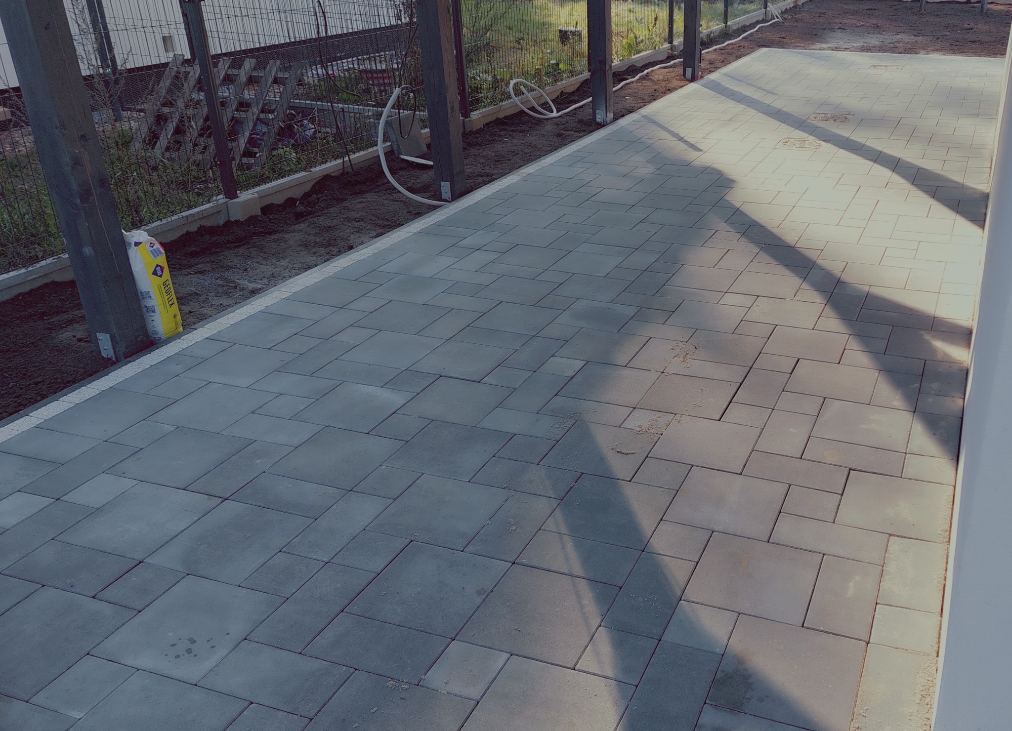 Ułożona szara kostka brukowa na posesji, widoczny worek z zaprawą, elementy ogrodzenia i cień. Kostka ułożona w regularny wzór, widoczne łączenia.