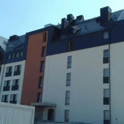 Wielopiętrowy budynek mieszkalny z dachem z blachy w kolorze grafitowym, częściowo wykończony, z widocznymi rusztowaniami i kontenerem budowlanym na pierwszym planie.