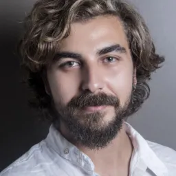 Portret młodego mężczyzny z brodą, kręconymi włosami i białą lnianą koszulą na szarym tle.