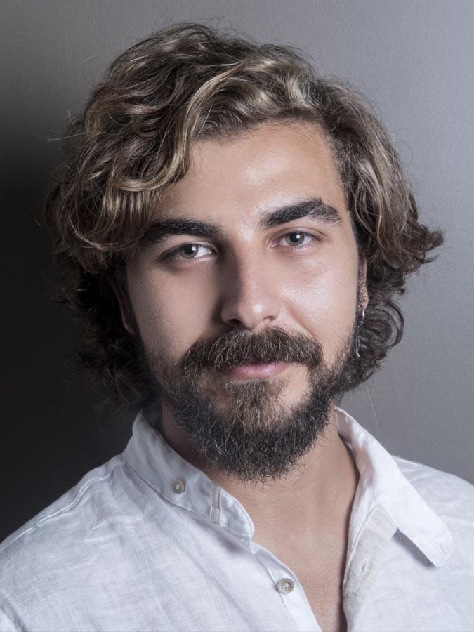 Portret młodego mężczyzny z brodą, kręconymi włosami i białą lnianą koszulą na szarym tle.