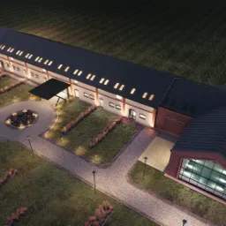 Wizualizacja z lotu ptaka budynku usługowego z czerwonej cegły z czarnym dachem, oświetlonym wejściem i zagospodarowanym terenem zielonym z lampami.
