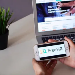 Smartfon z logo FreeHR trzymany na tle otwartego laptopa z widoczną stroną internetową agencji rekrutacyjnej, obok doniczka z zieloną trawą.