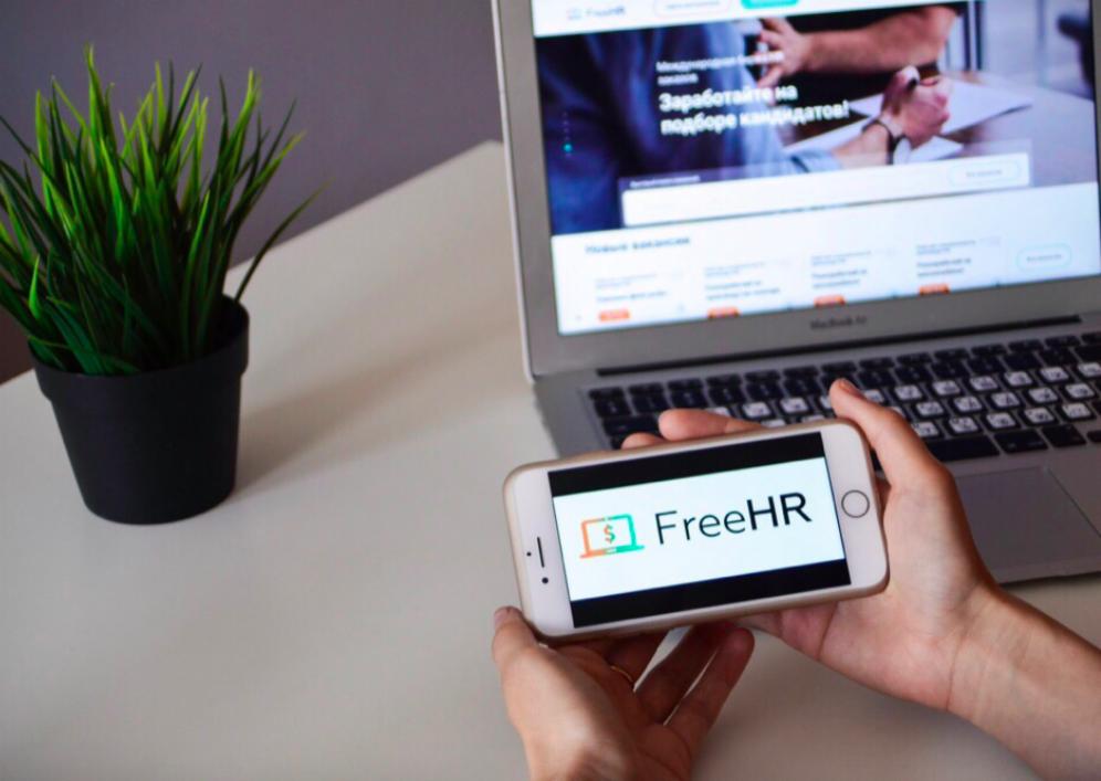 Smartfon z logo FreeHR trzymany na tle otwartego laptopa z widoczną stroną internetową agencji rekrutacyjnej, obok doniczka z zieloną trawą.