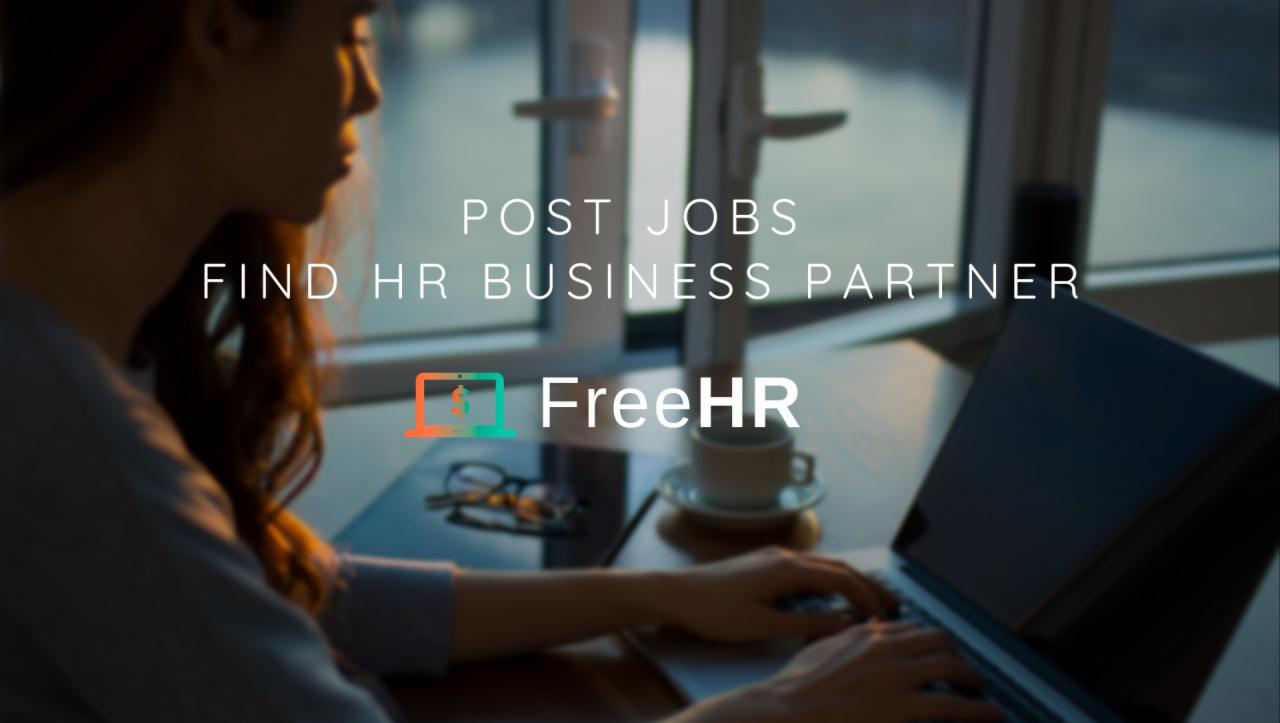 Kobieta pisze na laptopie przy biurku z logo FreeHR, okulary i filiżanka kawy obok, za oknem widok na miasto.