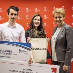 Trzy osoby pozują do zdjęcia z voucherem na 30 godzin doradztwa prawnego i torbą z napisem #KOCHAMŁÓDŹ na tle ściany z logo Uniwersytetu Łódzkiego.