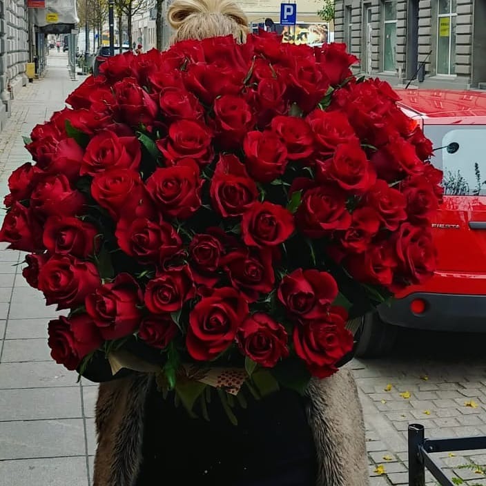 Ogromny bukiet stu czerwonych róż niesiony na tle miejskiego chodnika i czerwonego samochodu.