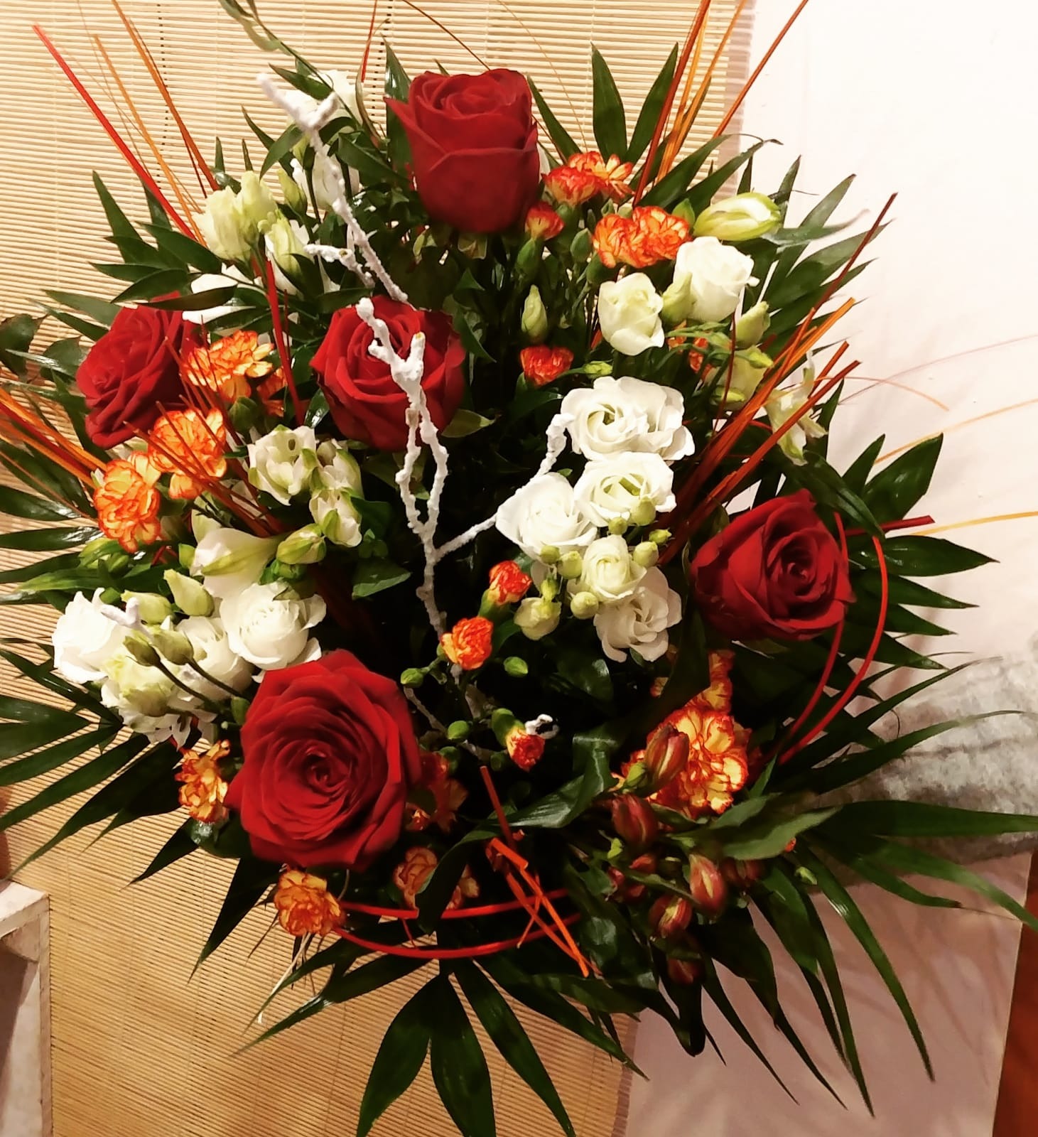 Kompozycja kwiatowa z czerwonych róż, białych i pomarańczowych goździków, zielonych liści palmowych i dekoracyjnych gałązek na tle bambusowej rolety.