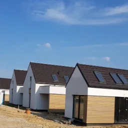 Rząd nowoczesnych, parterowych domów z drewnianą elewacją i ciemnymi dachami z oknami połaciowymi, usytuowanych na placu budowy pod błękitnym niebem.