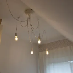 Nowoczesna lampa wisząca z pięcioma żarówkami na białych kablach, zamontowana na suficie w pokoju z oknem i tapetą w zygzaki.