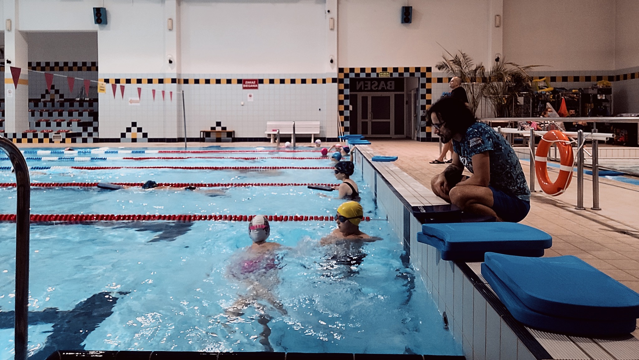Kryty basen z kilkoma osobami pływającymi po torach, ratownik w okularach siedzi na krawędzi basenu obok pomarańczowego koła ratunkowego