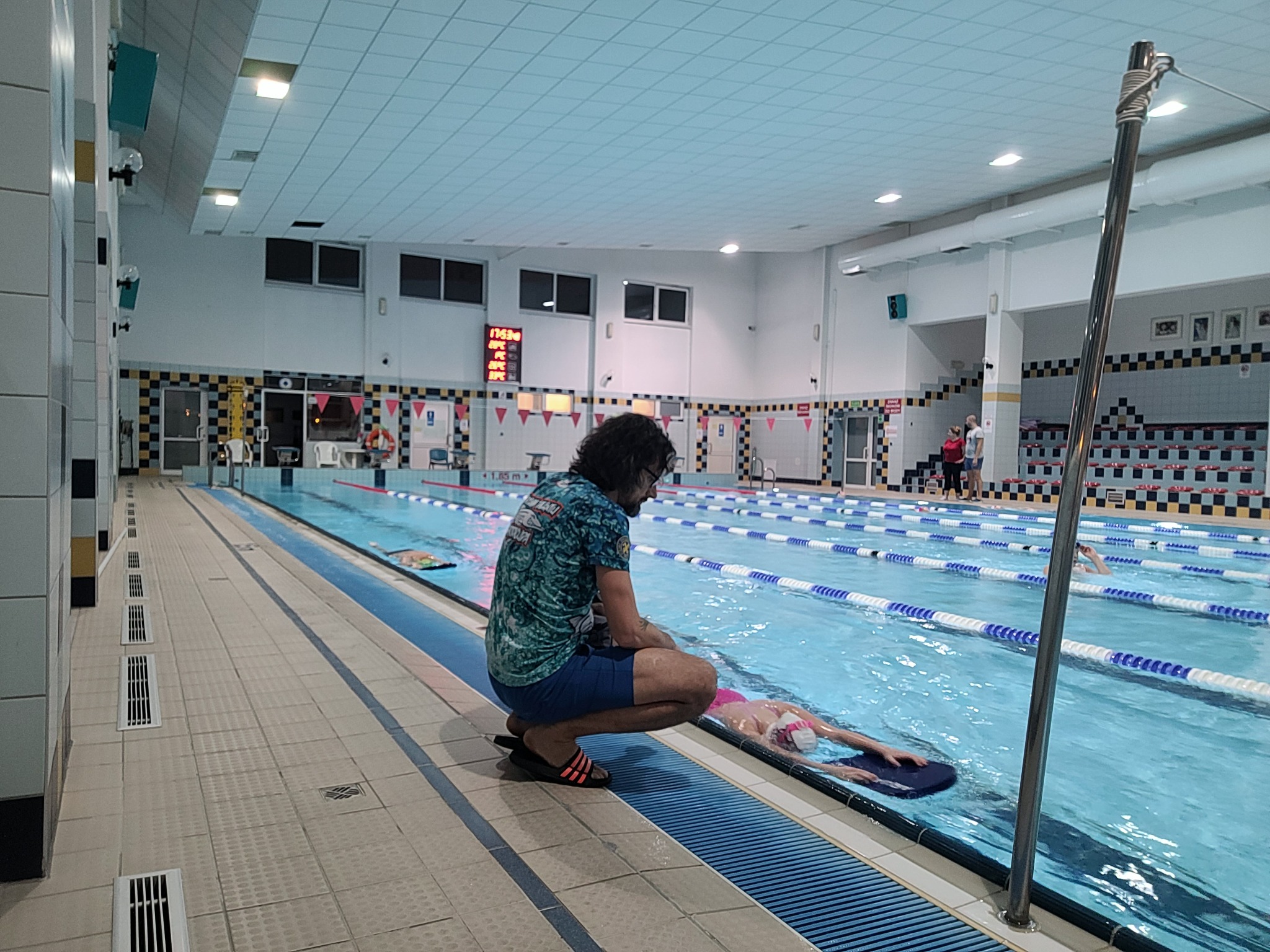 Instruktor w okularach kuca przy basenie, obserwując pływaczkę z deską w różowym czepku, na tle oświetlonego basenu z torami i trybunami.