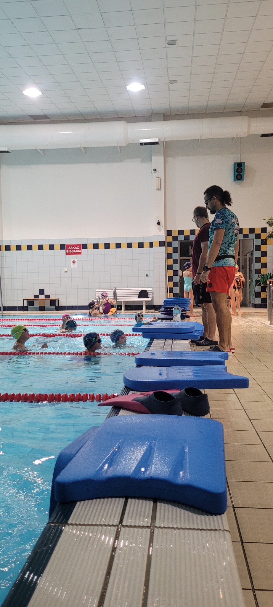 Grupa dzieci w czepkach pływackich w basenie podczas zajęć, instruktorzy stoją na brzegu. Na pierwszym planie niebieskie deski do pływania i płetwy.