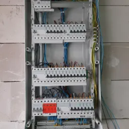 Szara skrzynka rozdzielcza z zainstalowanymi bezpiecznikami i przewodami, widoczna na tle ściany z białych bloczków. Zabezpieczenia SIMTEC z czerwonymi elementami. Przewody w kolorach niebieskim...