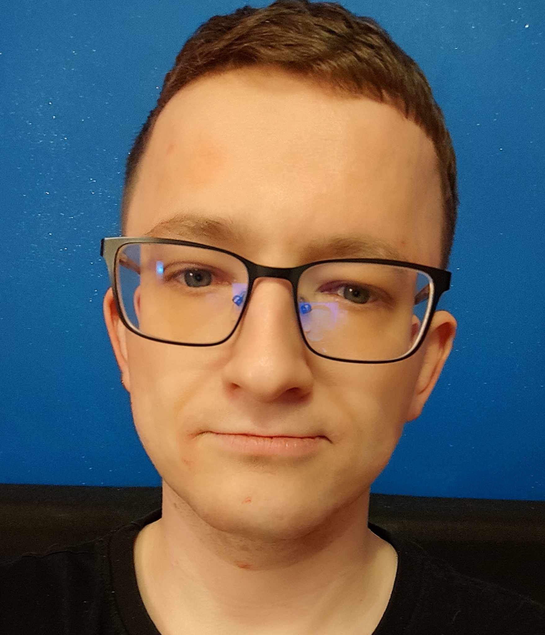 Portret młodego mężczyzny w okularach na tle błękitnej ściany z drobnymi białymi plamkami.