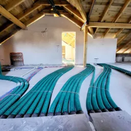 Instalacja rekuperacji w surowym stanie poddasza, widoczne zielone rury wentylacyjne prowadzone po podłodze w kierunku otworu drzwiowego, drewniana konstrukcja dachu w tle.