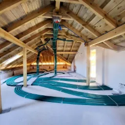 Zielone rury wentylacyjne ułożone na podłodze poddasza z drewnianą konstrukcją dachu w trakcie montażu.