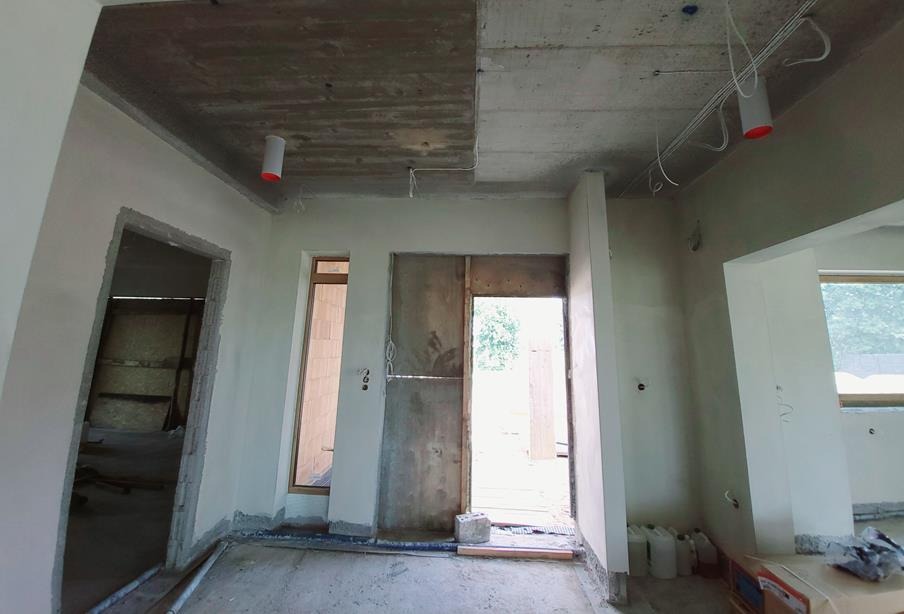Wnętrze w stanie surowym: widoczne instalacje elektryczne pod sufitem, niezabudowane otwory okienne i drzwiowe, ściany częściowo otynkowane, elementy systemu wentylacyjnego.