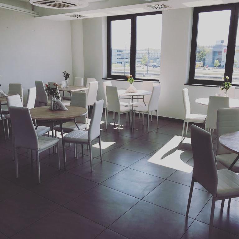 Jasne, przestronne wnętrze sali konferencyjnej z okrągłymi stołami, białymi krzesłami i minimalistycznymi dekoracjami kwiatowymi, oświetlone promieniami słońca wpadającymi przez okna.