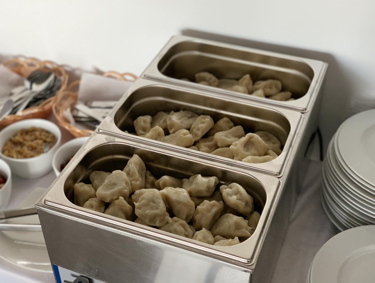 Trzy metalowe pojemniki GN wypełnione pierogami, ustawione jeden na drugim, obok stosu białych talerzy i koszyka ze sztućcami. Widoczne również miseczki z dodatkami.