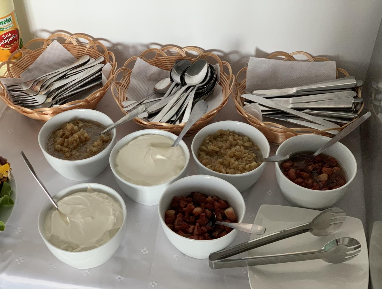 Bufet z przystawkami: sos jalapeño, koszyki ze sztućcami, miseczki z sosem czosnkowym, sosem tatarskim, skwarkami i sosem pomidorowym z boczkiem, część sałatki w tle.