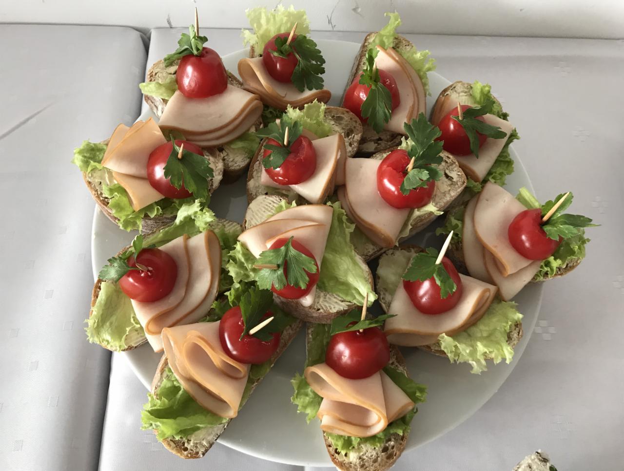 Dekoracyjne kanapeczki na ciemnym pieczywie z sałatą, szynką i pomidorkiem koktajlowym, ozdobione listkiem pietruszki, podane na białym talerzu.