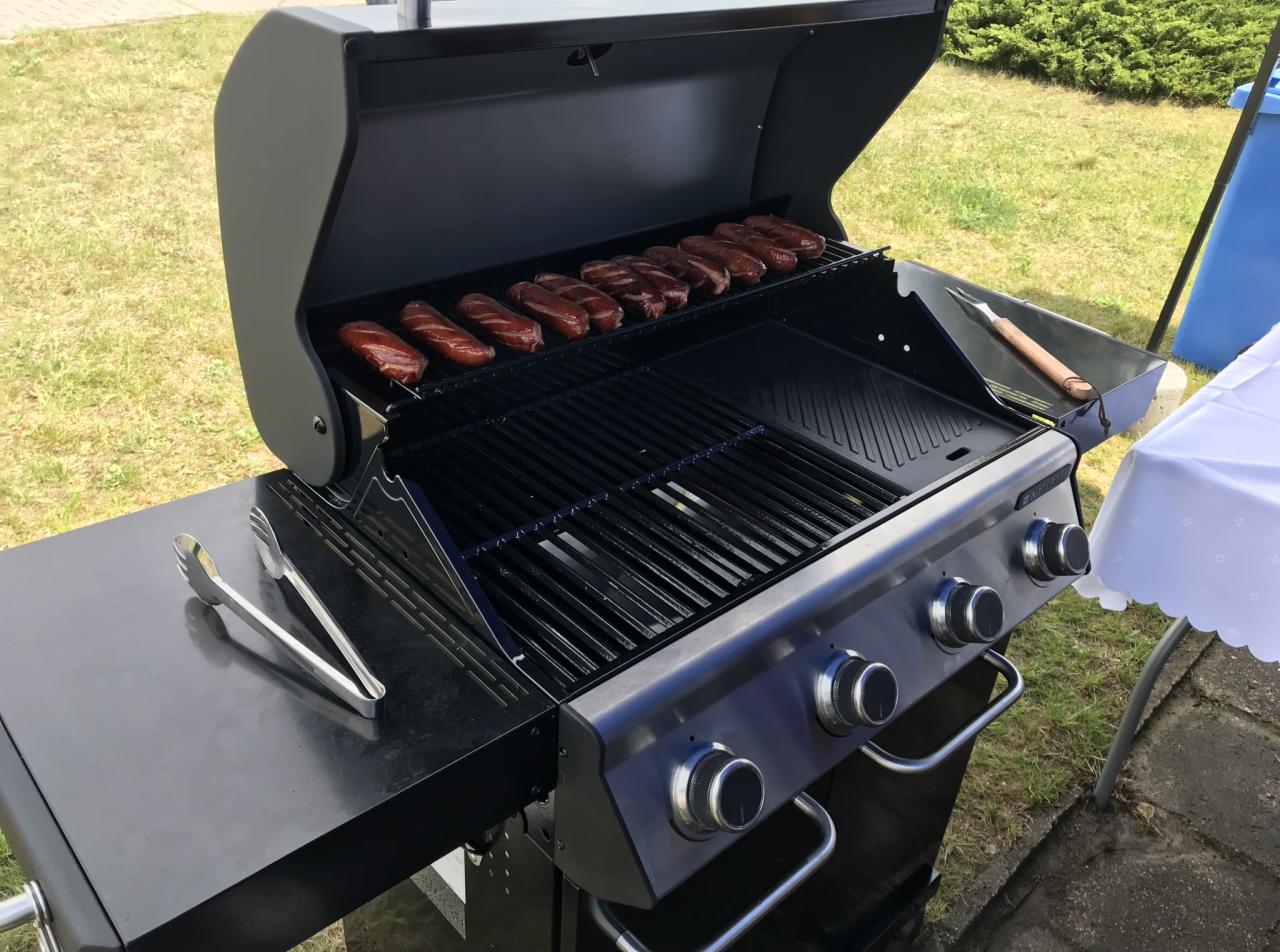 Grill gazowy z rzędem brązowiejących kiełbas na ruszcie, szczypce obok, tło trawnik i fragment stołu z białym obrusem.