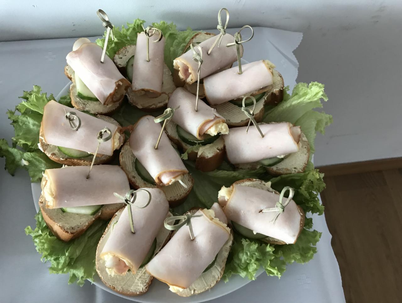 Dekoracyjnie ułożone kanapeczki z szynką, ogórkiem i sałatą na białym talerzu, zabezpieczone ozdobnymi wykałaczkami.