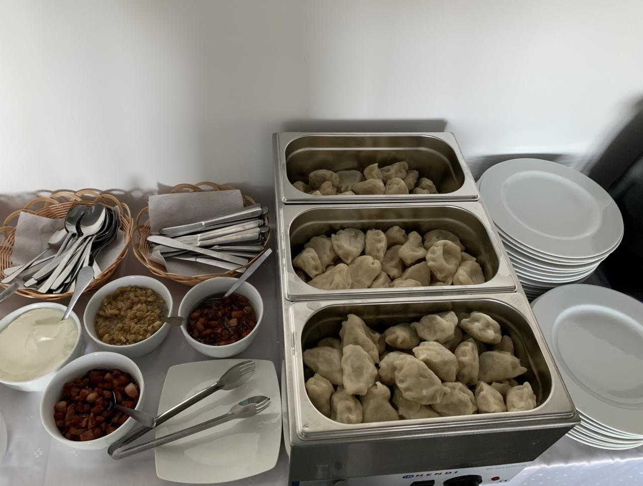 Bufet z pierogami w podgrzewanych pojemnikach, obok miseczki z sosem, śmietaną i skwarkami oraz stos białych talerzy i koszyczki ze sztućcami.