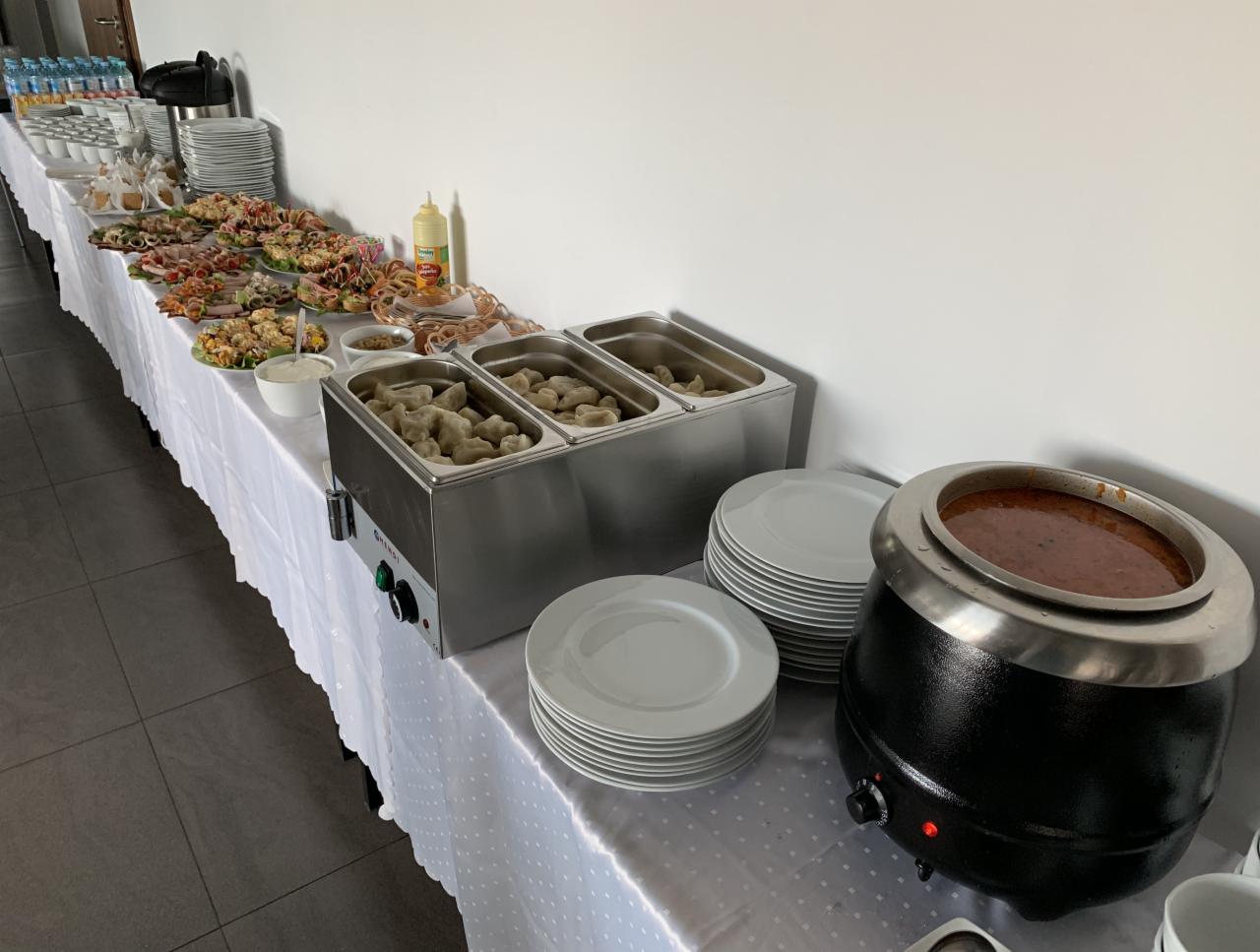 Stół z białym obrusem zastawiony różnorodnymi potrawami: sałatkami, wędlinami, pierogami w podgrzewaczach oraz zupą w czarnym kotle, przygotowanymi do serwowania.