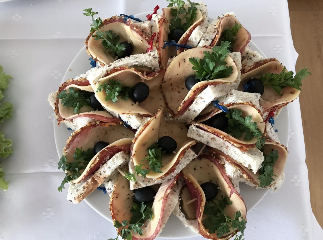 Dekoracyjnie ułożone na białym talerzu kanapki z jasnego pieczywa, szynki, sera, oliwek i zielonej pietruszki, spięte kolorowymi wykałaczkami.