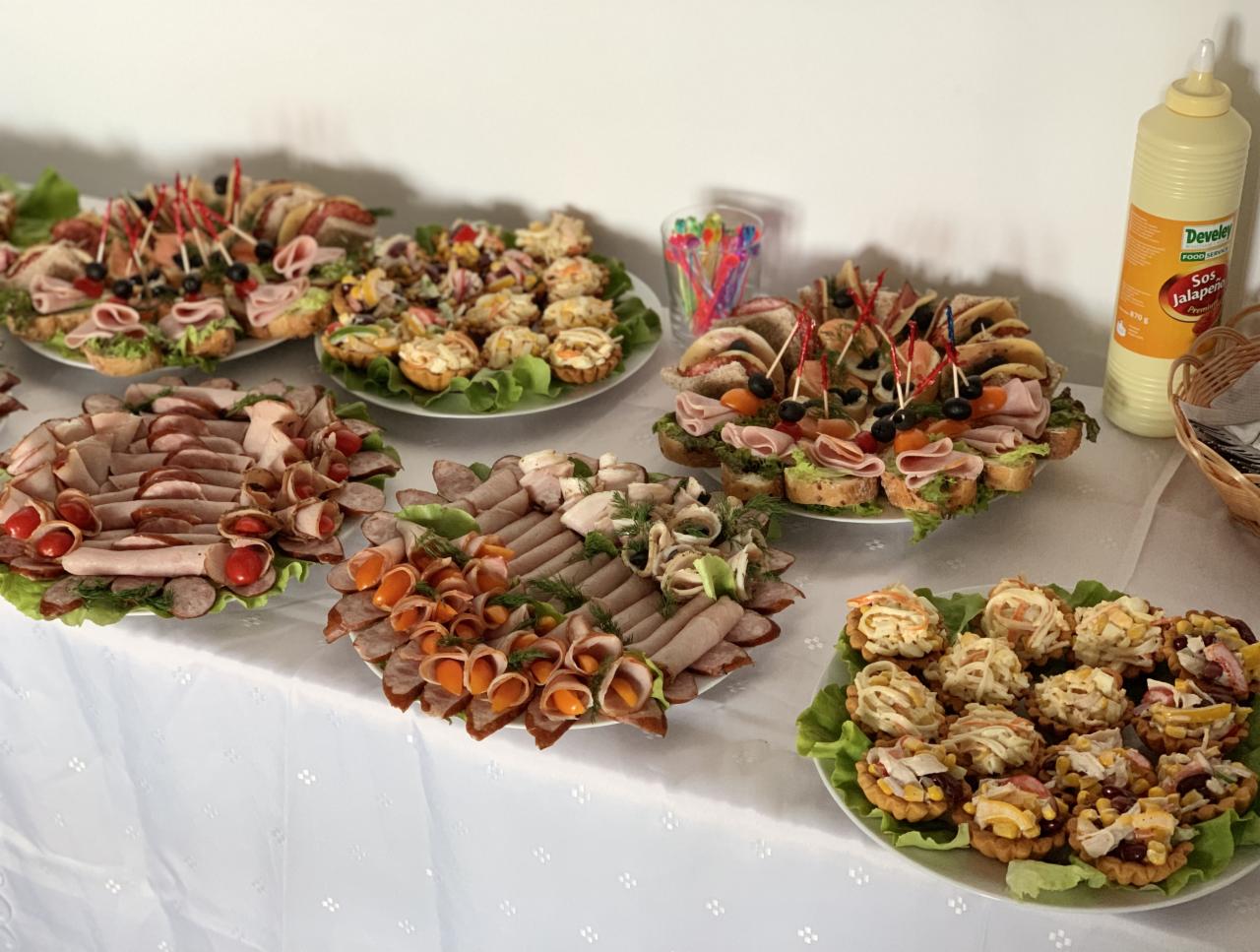 Stół z białym obrusem, na którym znajdują się talerze z różnorodnymi przekąskami: kanapki z szynką i oliwkami na wykałaczkach, tartaletki z sałatką, wędliny zwinięte w rulony z pomidorkami...